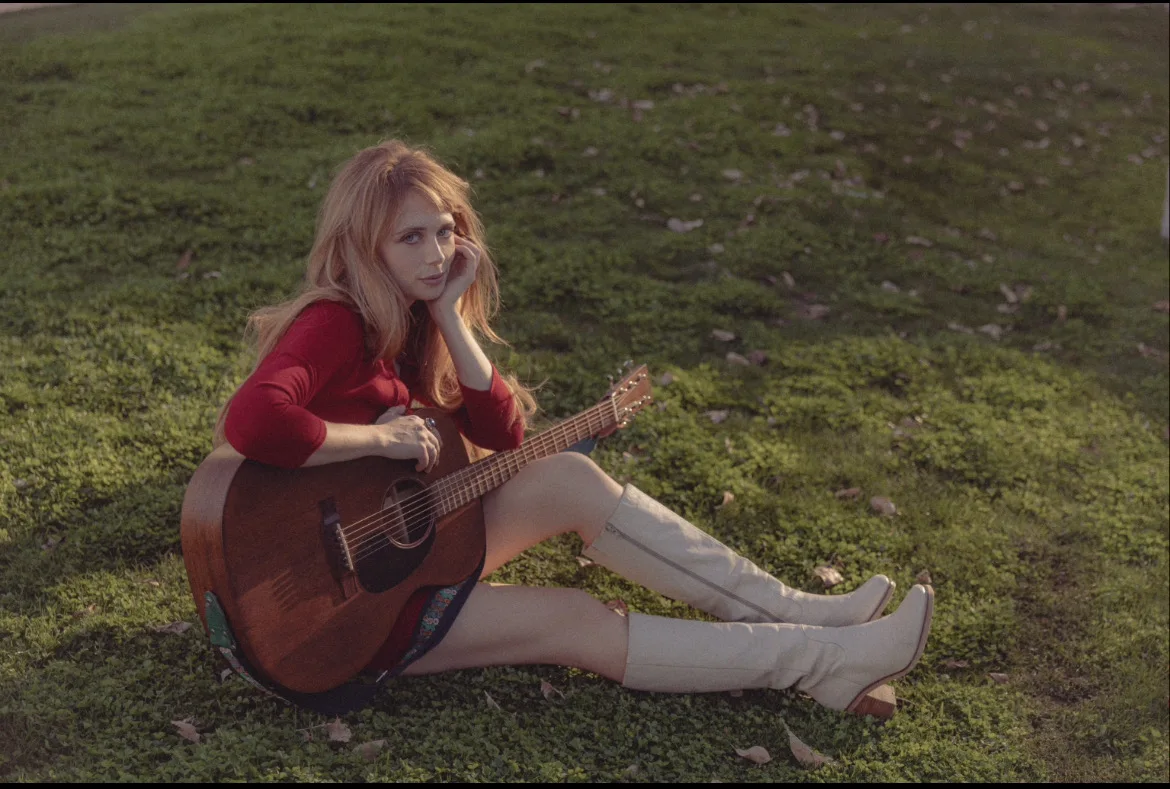 Autumn Cooper sits on grass with an acoustic guitar.