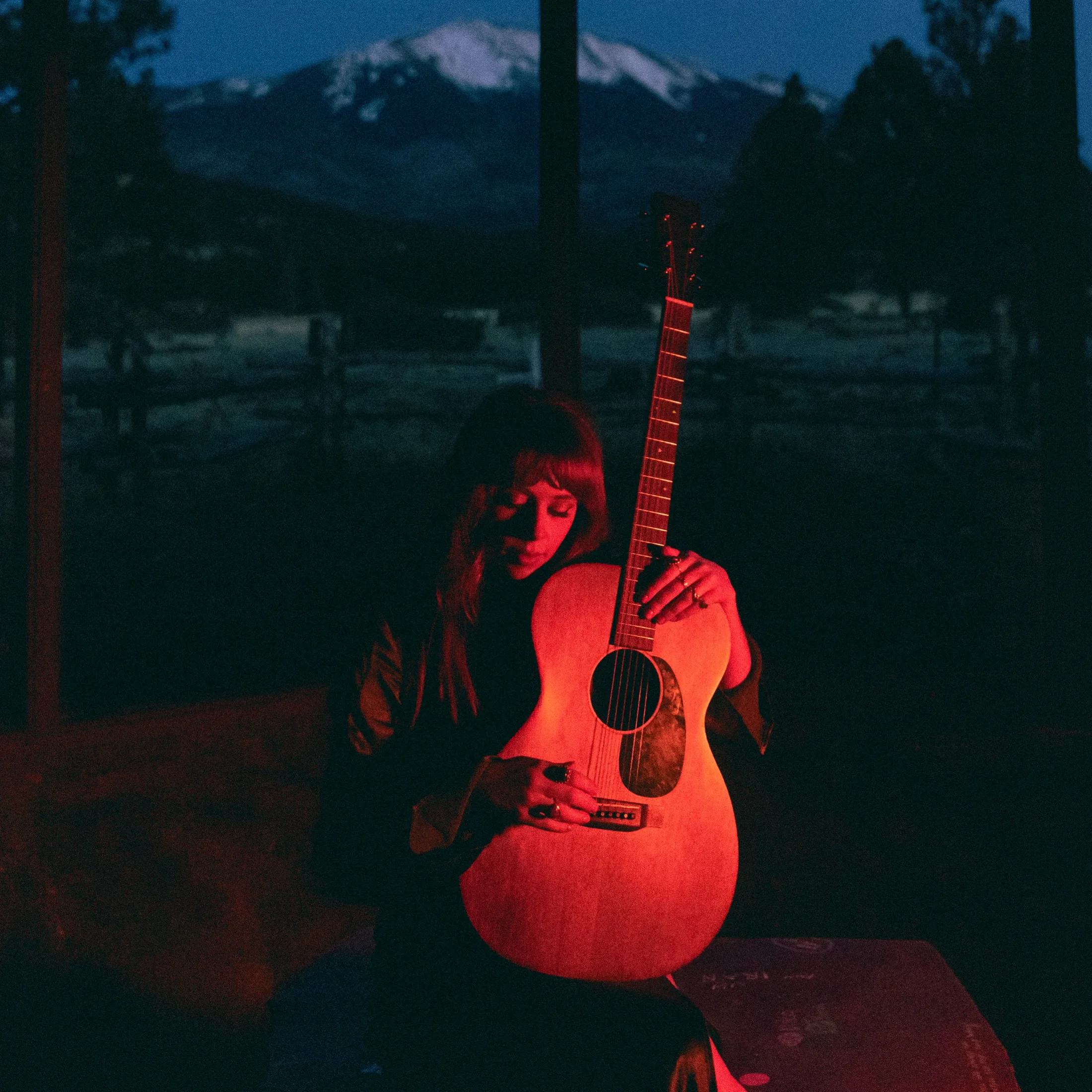Miss Autumn holds an acoustic guitar in red light with mountains visible through a window.